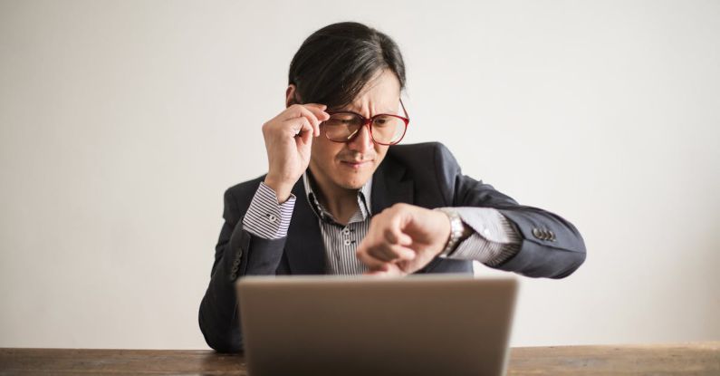 Meeting Deadlines - Young frowning man in suit and glasses looking at wristwatch while waiting for appointment sitting at desk with laptop